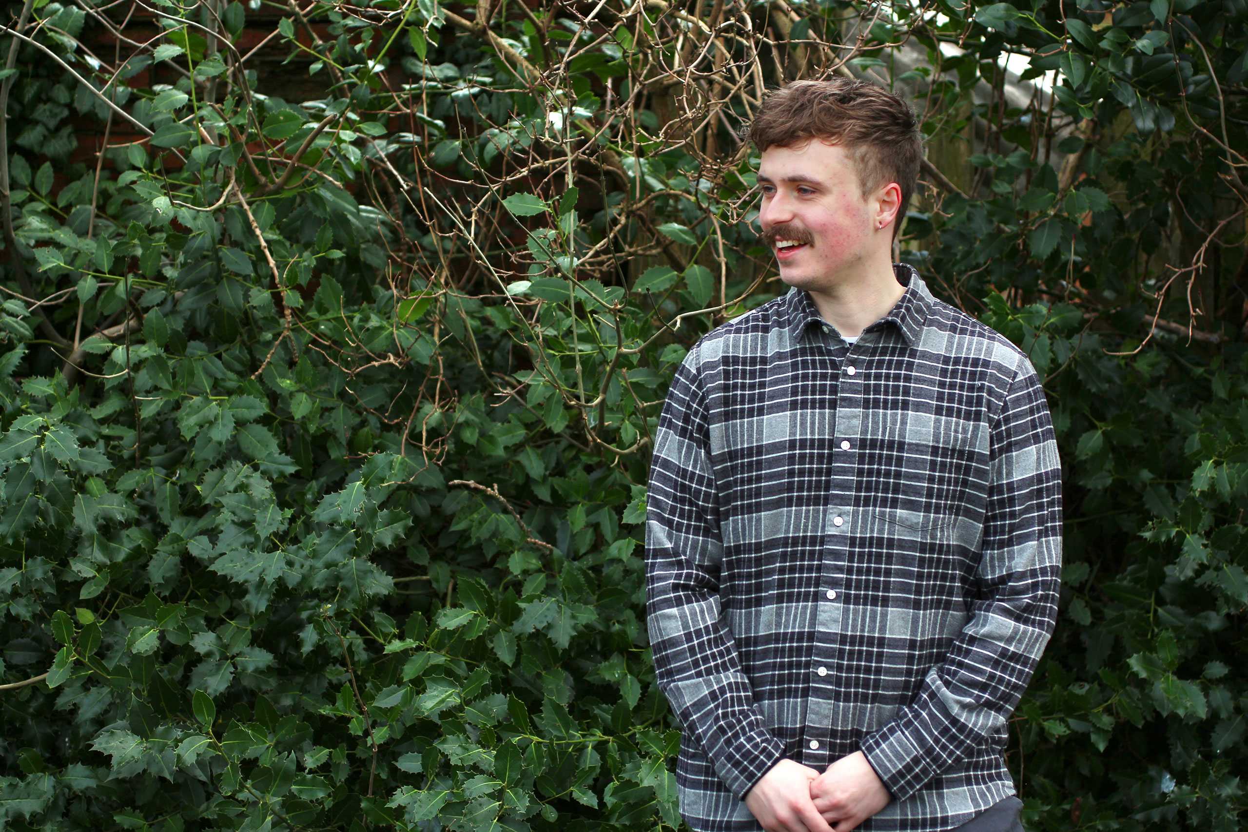 Toby Lyons in a shirt stood in front of a leafy bush